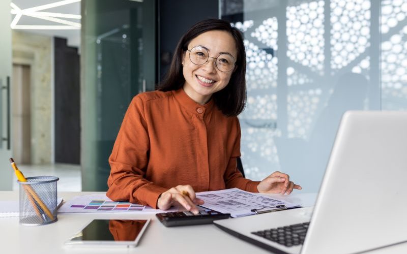 Portrait of young Asian successful accountant working on paperwork with calculator inside office